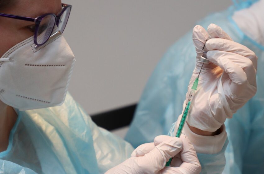 pielęgniarka w fartuchu ochronnym i masce pobiera strzykawką szczepionkę