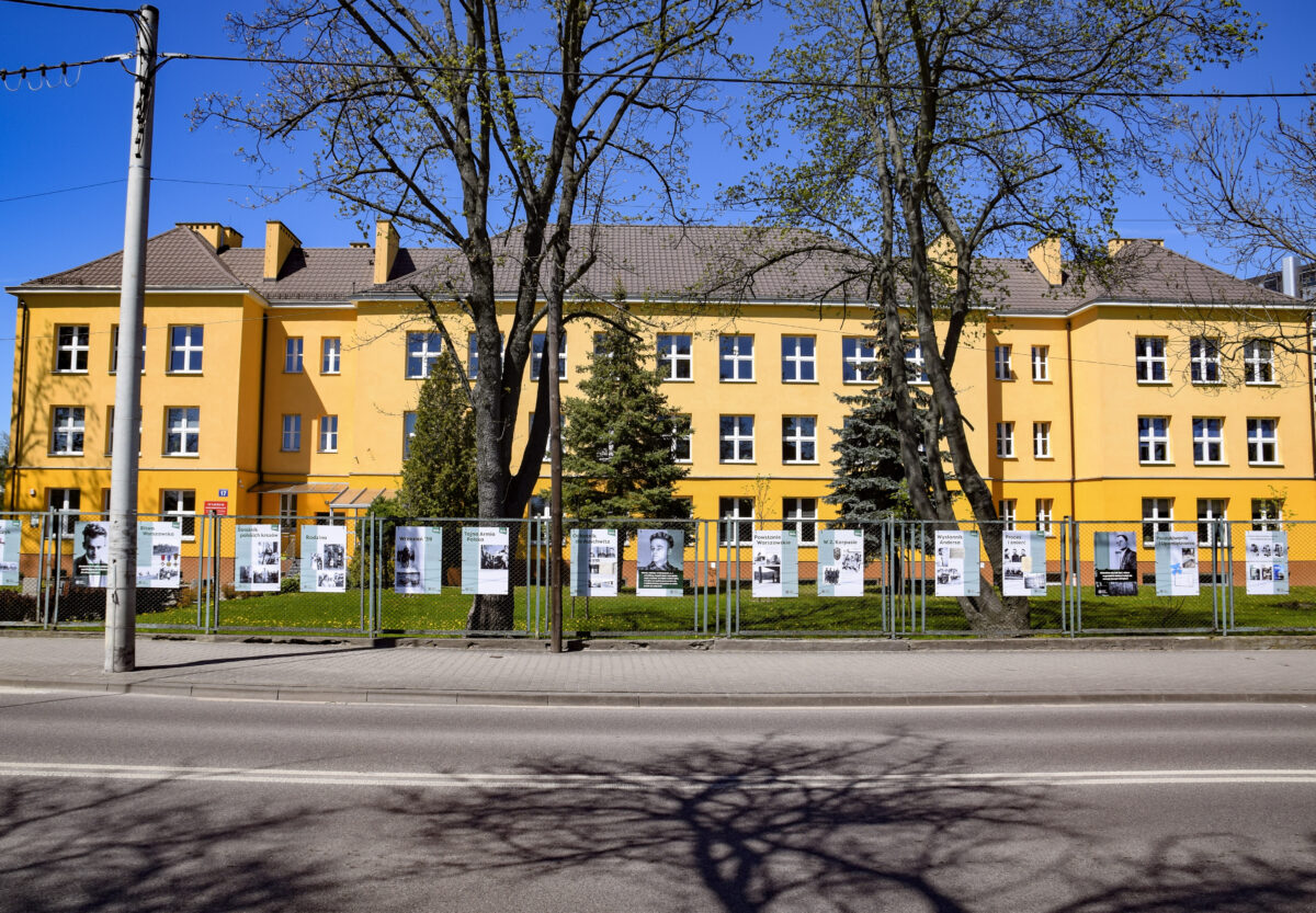 plansze na ogrodzeniu, w tle żółty budynek szkoły