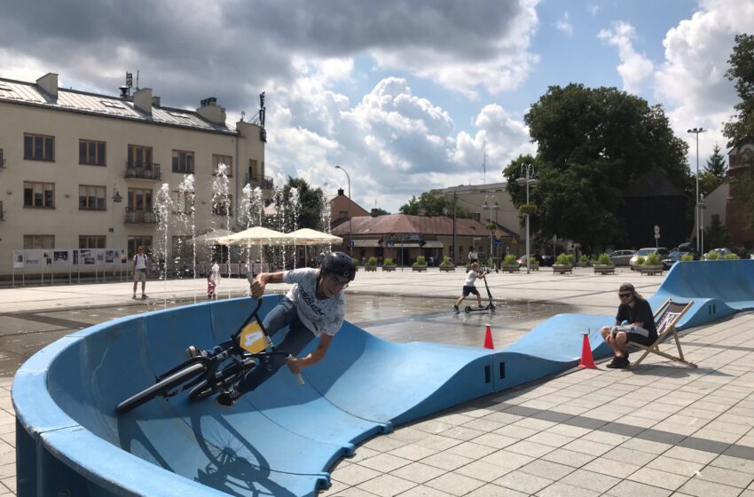 rowerzysta na torze, w tle dziecko na hulajnodze, fontanna