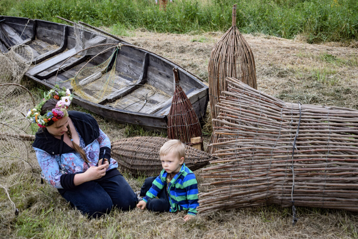 Mama z dzieckiem na tle drewnianej łodzi i wiklinowej wirażki do połowu ryb