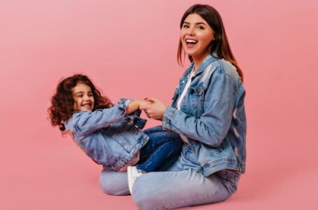 Cheerful caucasian woman playing with daughter. Studio shot of preteen curly child sitting with mom on pink background.