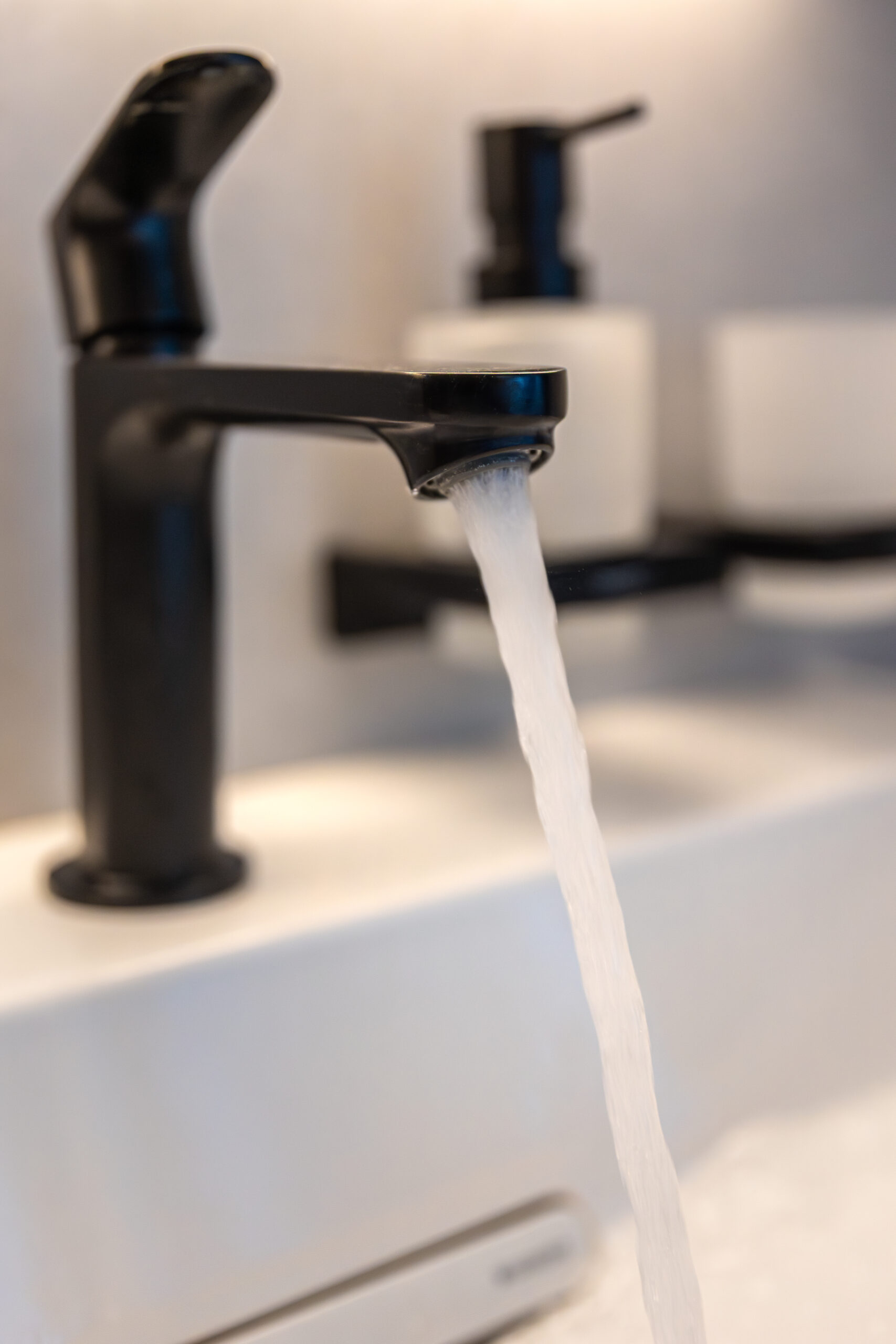 Vertical view of a matte faucet with an aerated foamy water stream into a white basin. Warm light and clean shapes create a spa mood.