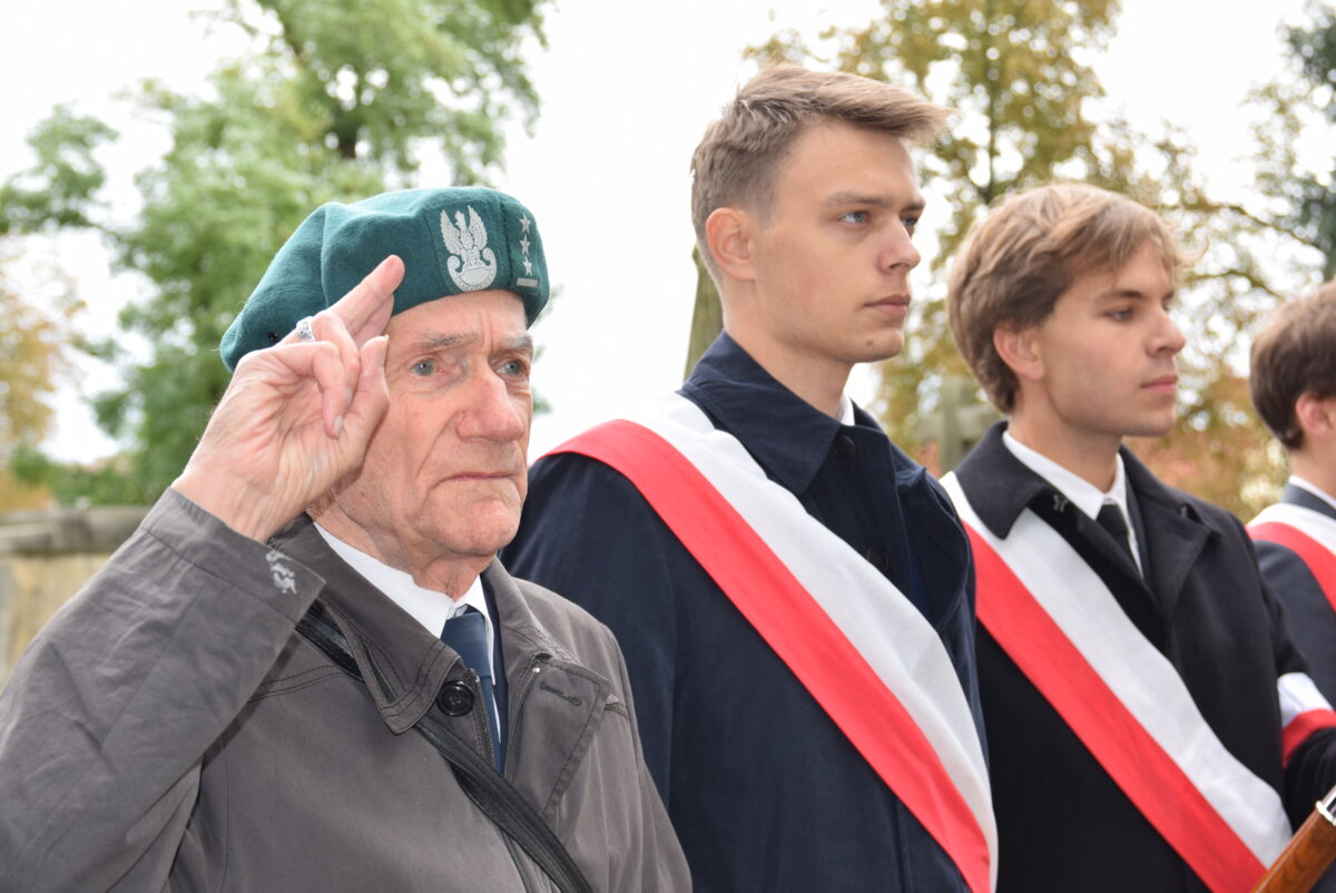 W. Sieradzki salutuje, obok 2 młodzi ludzie przepasani biało-czerwonymi szrfami